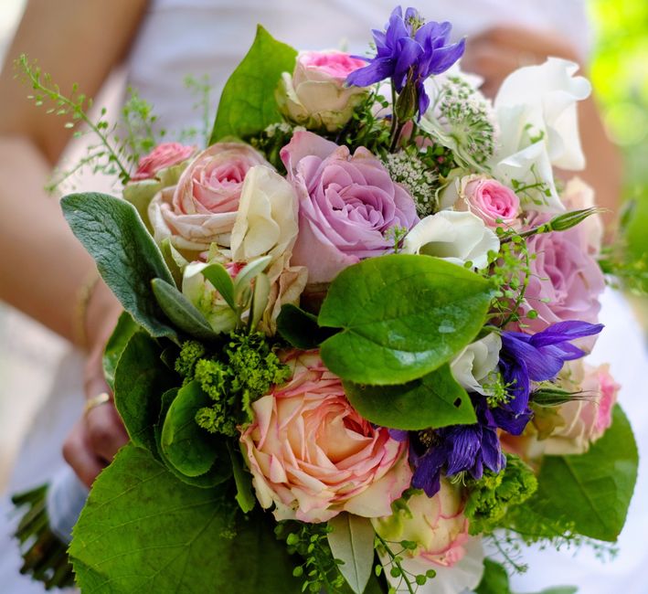 A hand-held bouquet featuring a combination of soft pink and white roses, accented by vivid purple irises and delicate white baby's breath. The arrangement is lush with large, glossy green leaves and 
