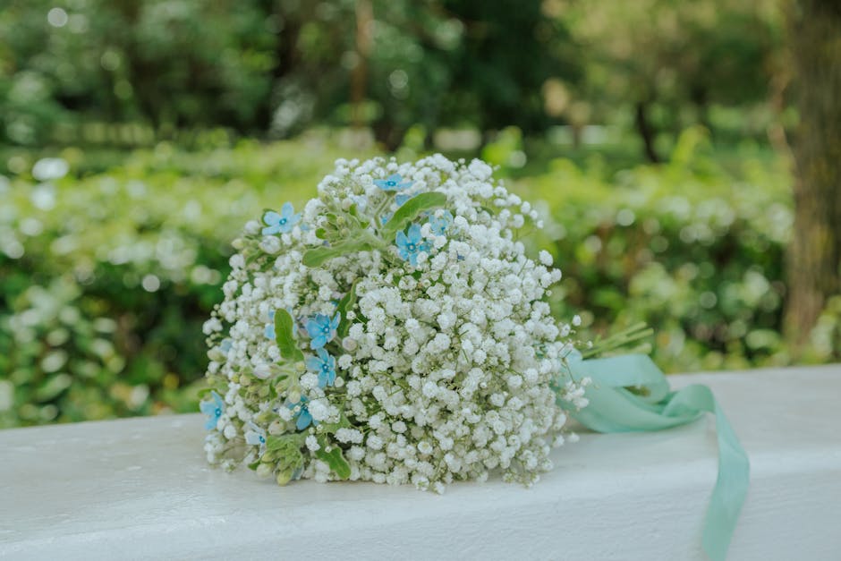 A delicate bouquet of small white gypsophila flowers arranged in a rounded, dome shape with subtle hints of light blue flowers interspersed among the white blooms. The bouquet is wrapped with a soft, 