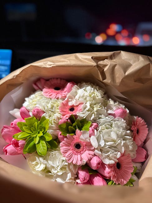 A bouquet of fresh flowers wrapped in brown paper, featuring pink gerbera daisies with dark centers, white hydrangeas with rounded clusters of petals, and green foliage accents. The arrangement is hel