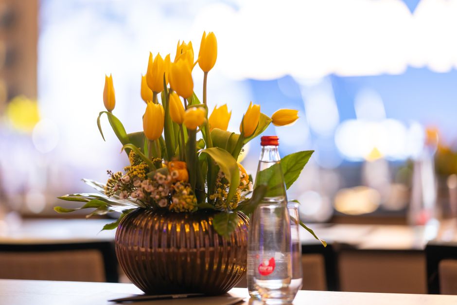 A floral arrangement featuring bright yellow tulips with closed buds and some partially open blooms, arranged in a dark, ribbed glass vase. The bouquet is accented with small, delicate pink and white 