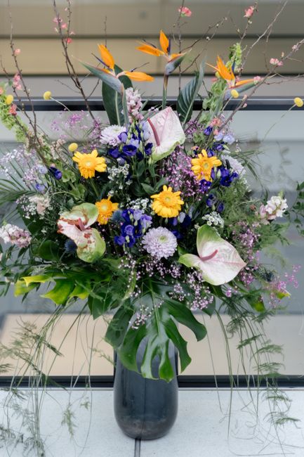 A tall floral arrangement in a dark, cylindrical vase featuring a variety of fresh flowers including large white calla lilies with pink streaks, bright yellow daisies, and vivid blue and purple blooms